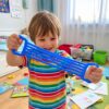 Bandă elastică din cauciuc, antistres, albastră 2 Bandă elastică din cauciuc, băiat într-un tricou în dungi colorate care întinde banda albastră cu ambele mâini, într-o cameră de joacă.